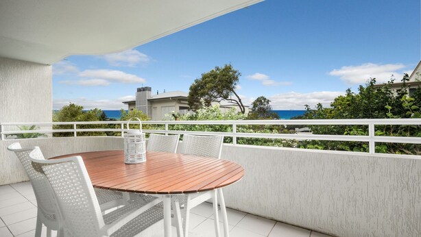 A round wooden table with white chairs on a balcony with a white railing, overlooking a coastal area with buildings and trees. - Cote D'Azur 4 Sunshine Beach (Sunshine Beach)