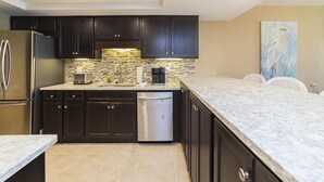 A modern kitchen with dark cabinets, a stainless steel refrigerator, a dishwasher, and a marble countertop.