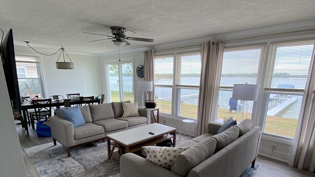 Ein geräumiges Wohnzimmer mit einer großen Couch, einem Couchtisch und einem Essbereich mit Tisch und Stühlen. Große Fenster mit Jalousien, ein Deckenventilator und Blick auf ein Gewässer.