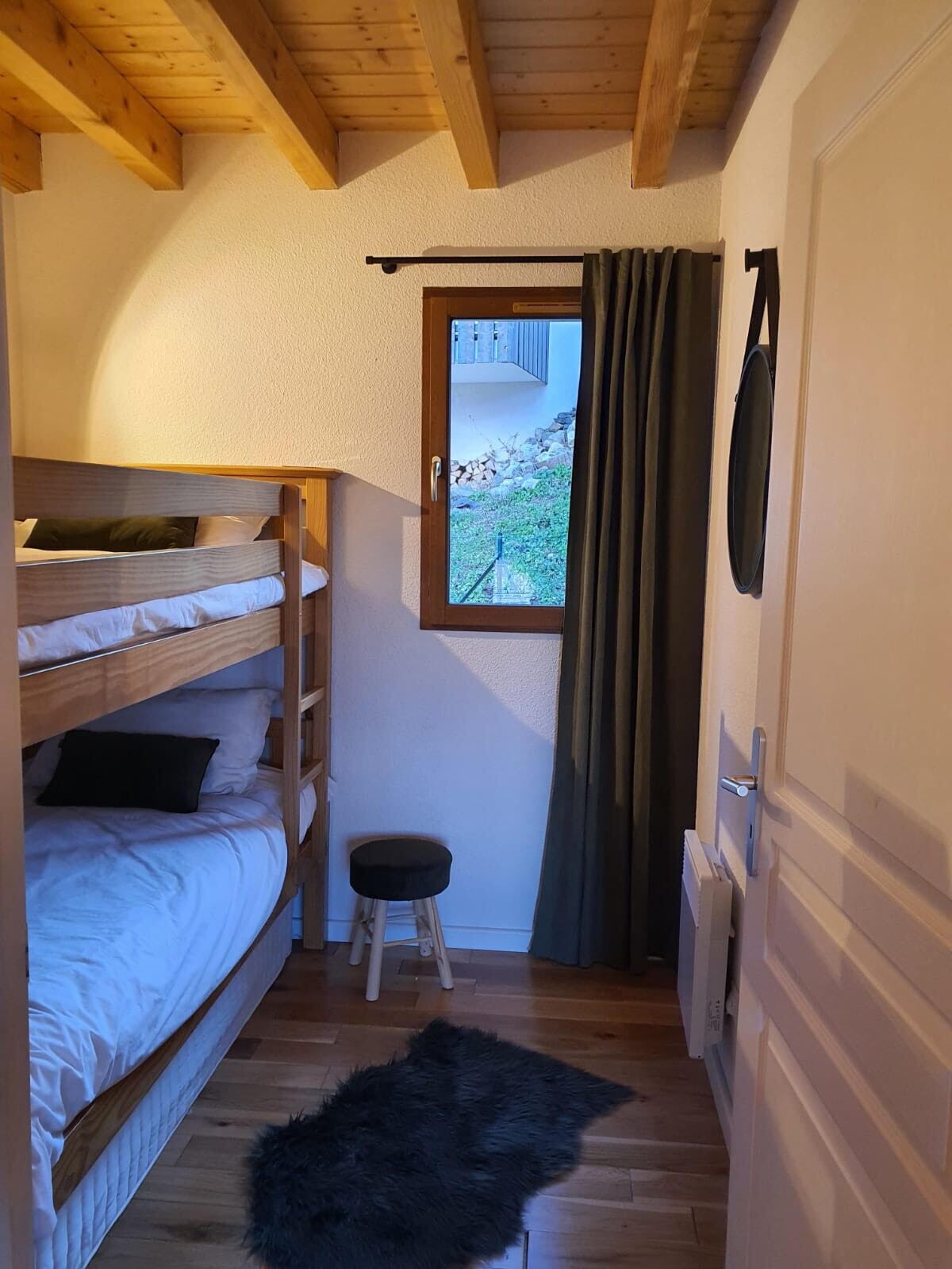Een kleine kamer met een stapelbed, een klein krukje, een raam met gordijnen en een radiator.