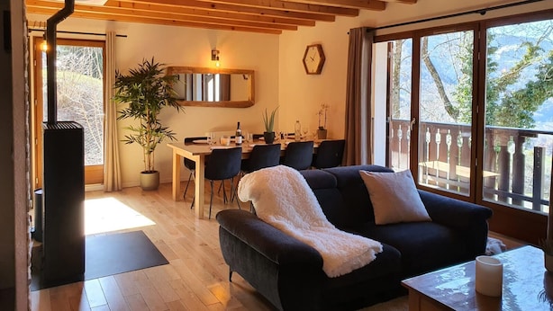 Ein gemütliches Wohnzimmer mit einer dunkelgrauen Couch, einem Essbereich mit einem Holztisch und Stühlen, und großen Fenstern mit Blick ins Freie.