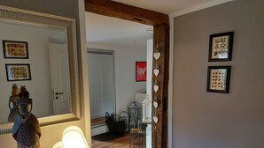 Un couloir avec un grand miroir, une lampe et une porte en bois. Les murs sont ornés de tableaux encadrés et un tapis recouvre le sol.