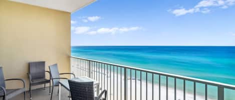 A balcony with wicker chairs and a table, overlooking a beach with clear turquoise water and a sandy shore.