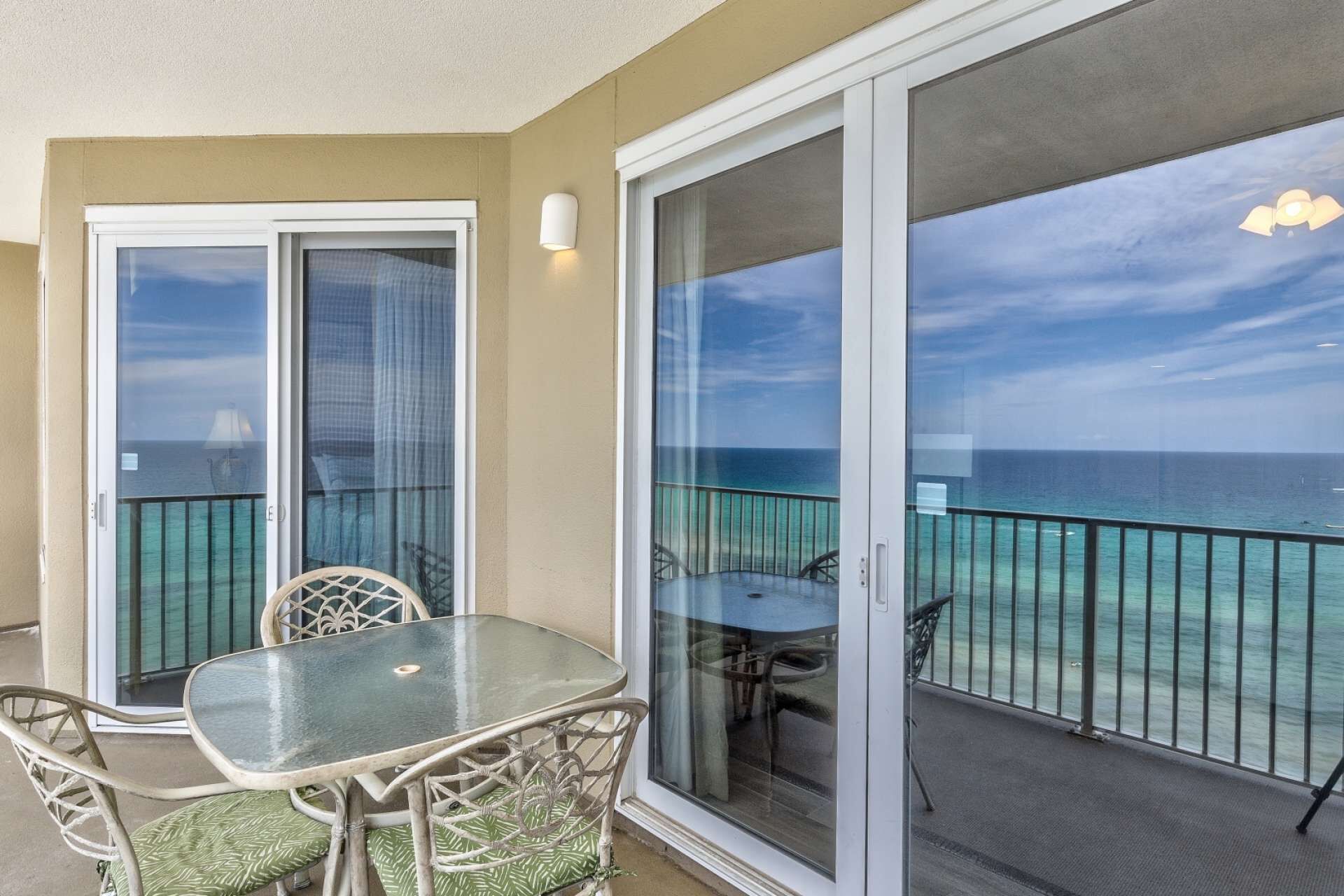 A balcony with a glass door, a table, and chairs, overlooking a turquoise sea.