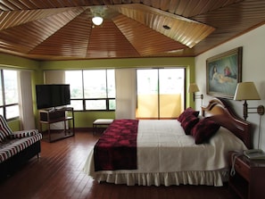 A hotel room with a wooden ceiling, a bed with a white and red bedspread, a television, a desk with a chair, and a painting on the wall. - Hotel Don Lolo (Villavicencio)