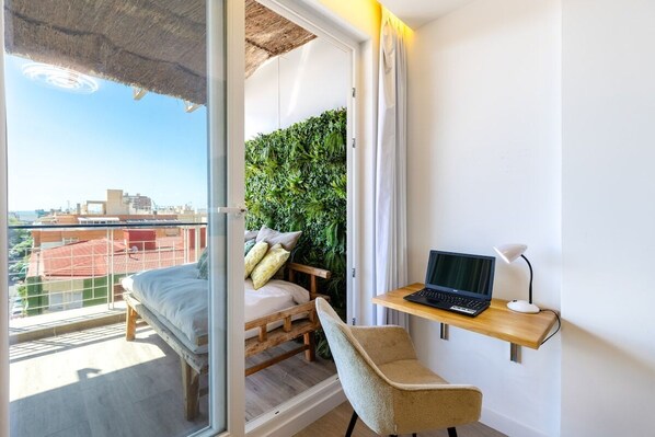 A bedroom with a bed, a desk with a laptop, a chair, a green wall, and a view of buildings.