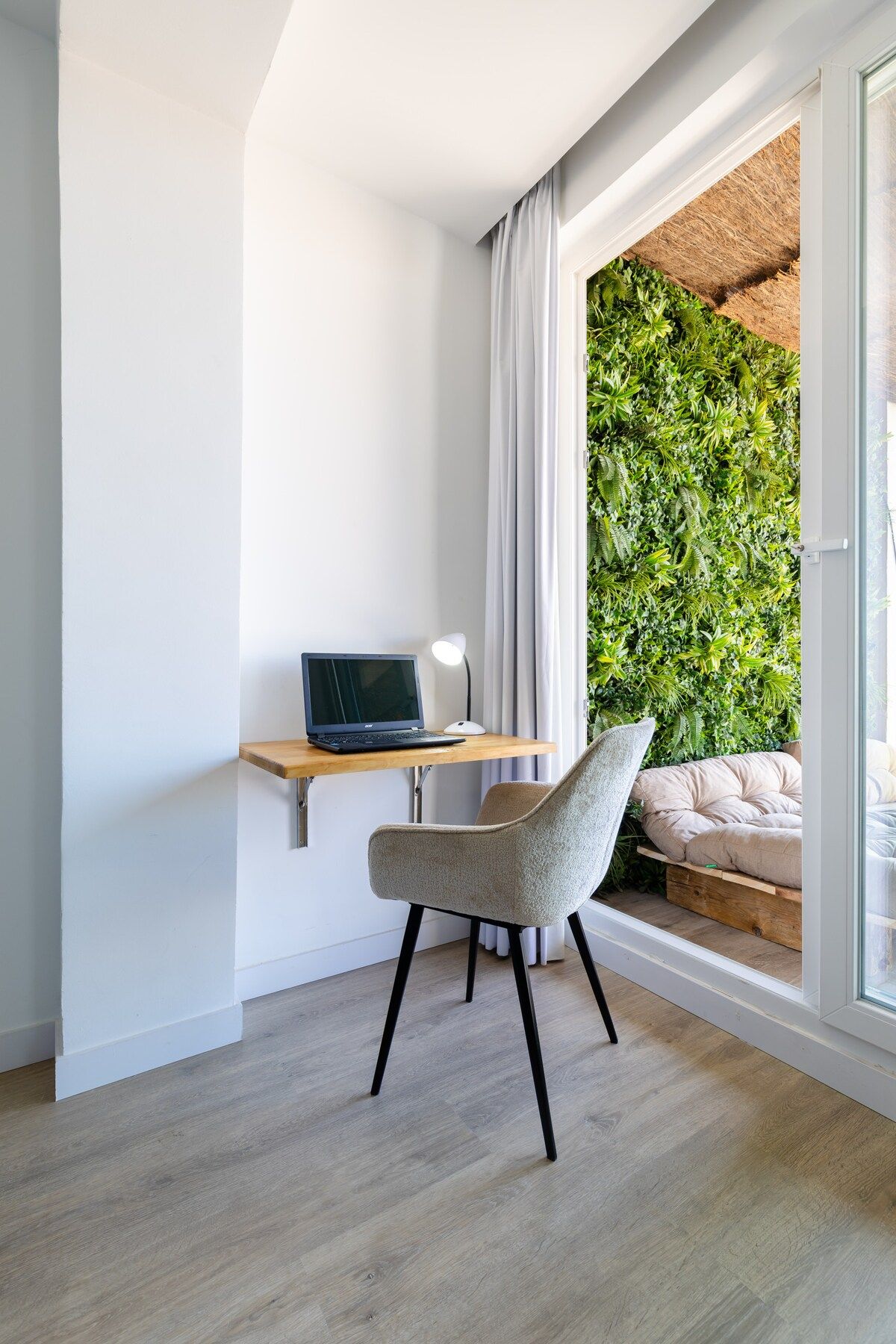 A modern room with a wooden desk, a laptop, a chair, and a large window with a view of a green wall and a bed.