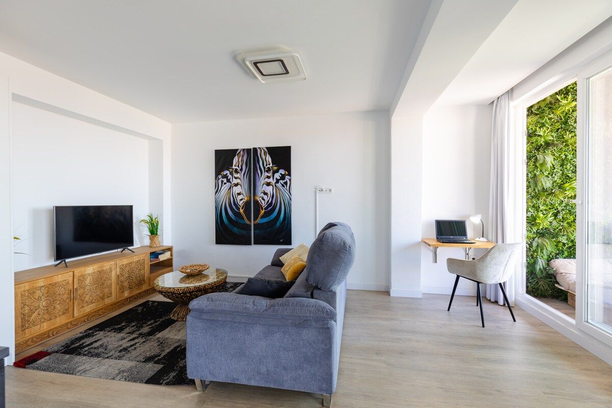 A modern living room with a grey sofa, a flat-screen TV, a wooden cabinet, and a large window with a view of greenery.