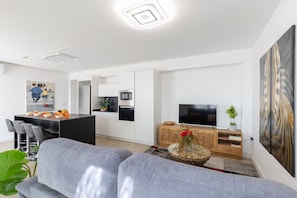 Ein modernes Wohnzimmer mit einer grauen Couch, einem Essbereich mit einer schwarzen Kücheninsel, einem Fernseher auf einem Holztisch und einem Gemälde an der Wand.