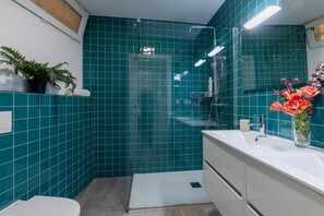 Ein Badezimmer mit blaustichigen Fliesen, einer Dusche, einem Waschbecken mit Spiegel und einer Topfpflanze.