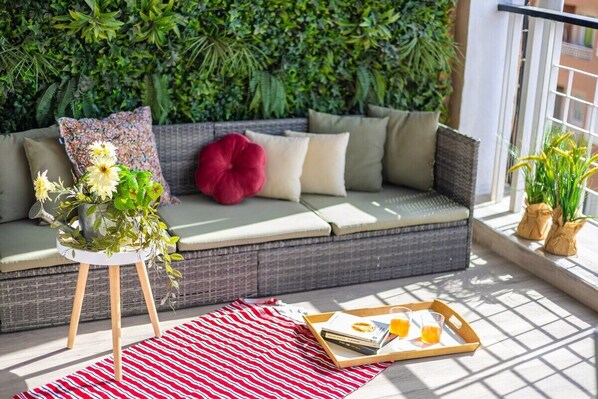 Ein rattangeflochtenes Sofa mit Kissen, ein rotes Kopfkissen, ein weißer Beistelltisch mit Pflanze und ein Tablett mit Büchern und Gläsern auf einem rot-weißen Streifen Teppich.