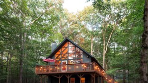 A cabin with a large glass front, wooden deck, and a staircase leading to a balcony.