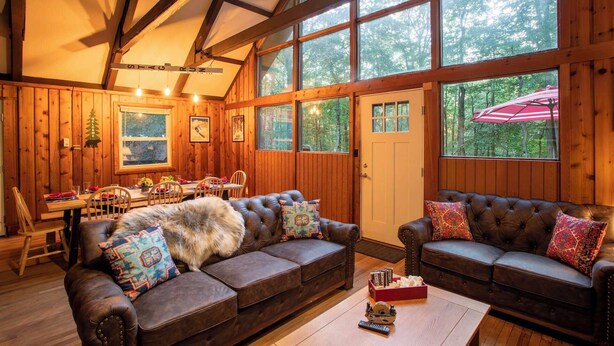 A cozy cabin interior with a dining area, two leather sofas, and large windows.
