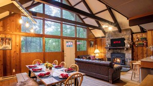 A cozy cabin interior with a dining area, sofa, fireplace, and large windows.