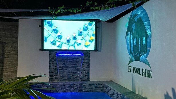 A swimming pool area with a waterfall feature, a mounted TV, and a mural of a house labeled "EZ POOL PARK". - Quite Suburbs of Bridgeport  (Portmore)