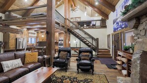A spacious interior with a high ceiling, a central staircase, leather armchairs, a wooden bench, and a stone fireplace.