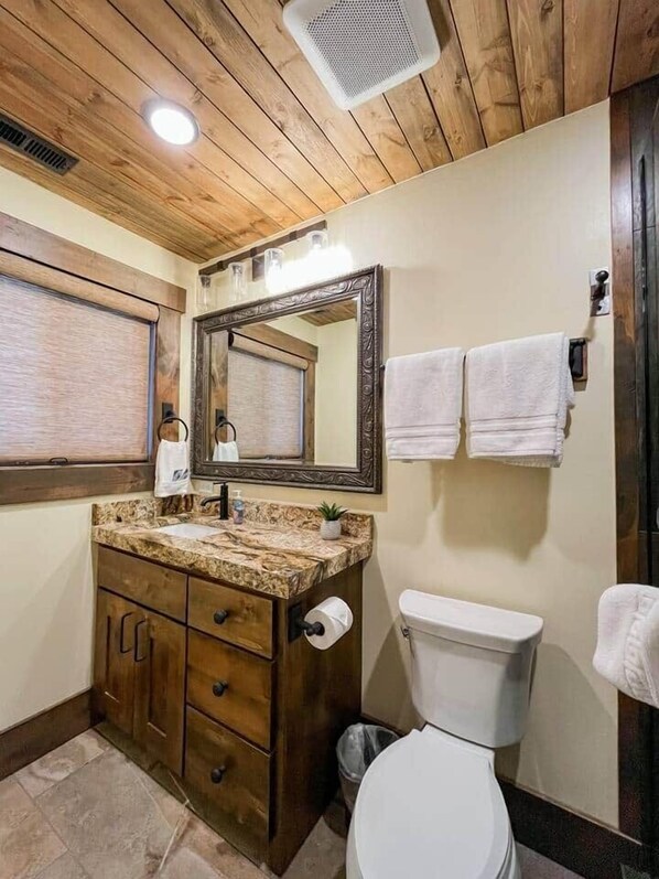 A bathroom with a wooden ceiling, a sink with a marble countertop, a toilet, and a mirror with a decorative frame.