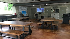 Un amplio espacio interior con varias mesas y bancos de madera, una pantalla de televisión mostrando un noticiero, y un cartel que anuncia cabañas de camping y pesca.