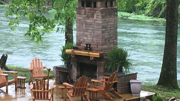 Una chimenea de piedra con chimenea, rodeada de sillas Adirondack de madera y una mesa, ubicada en una terraza junto a un río.