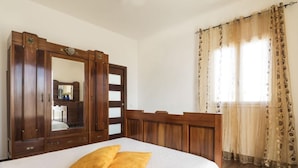 A bedroom with a large bed, a wooden wardrobe with a mirror, and a window with patterned curtains.