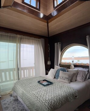 Une chambre avec un grand lit, une vue sur l’eau par la fenêtre et un plafond en bois.