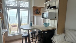 A compact kitchen with a central island, bar stools, a microwave, and a refrigerator. There is a window with a view of the sea, and a bed with a striped pillow. - Cozy Quiet Island Light House on Water Front (Fairgrove)