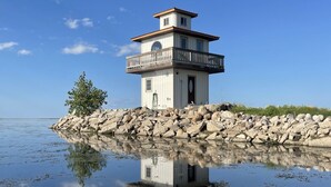A multi-story lighthouse with a balcony, situated on a rocky shore with calm water reflecting the structure. - Cozy Quiet Island Light House on Water Front (Fairgrove)
