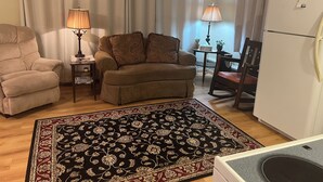 Ein Wohnzimmer mit Sofa, Sessel und Teppich. Auf einem Beistelltisch steht eine Lampe und in der Ecke steht ein Kühlschrank.