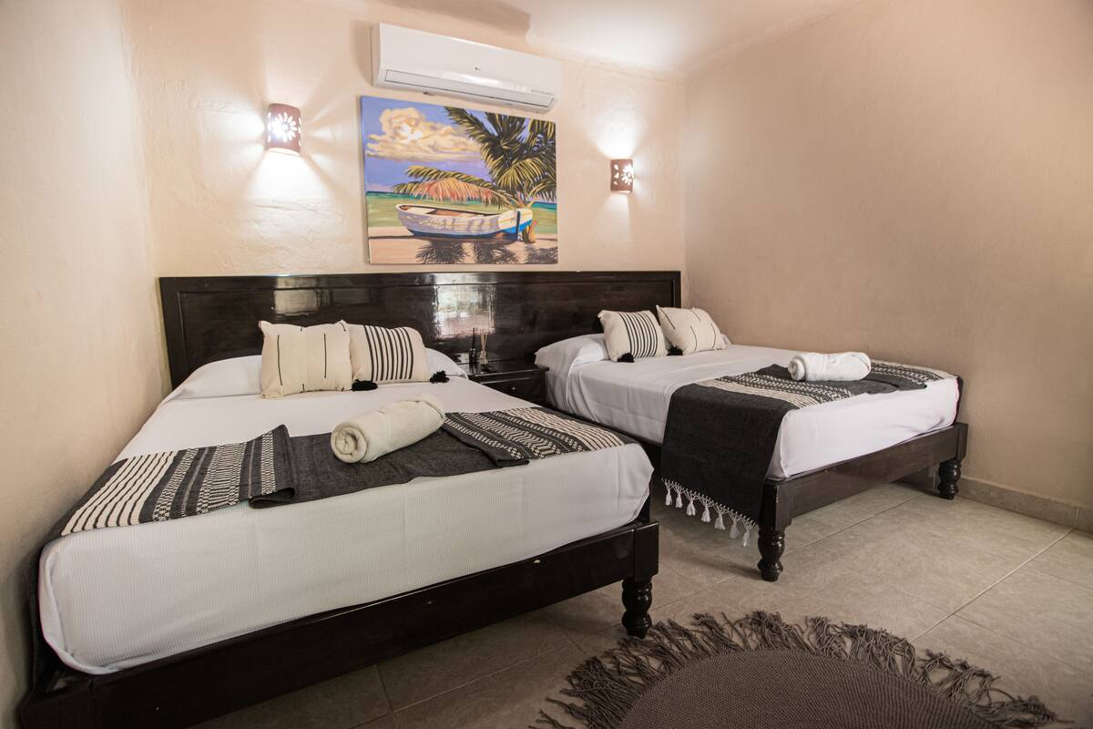Hotelkamer met twee bedden, elk voorzien van witte lakens, gestreepte kussens en een donker houten hoofdbord. Boven de bedden hangt een schilderij van een boot, en aan de muur is een airconditioner bevestigd.