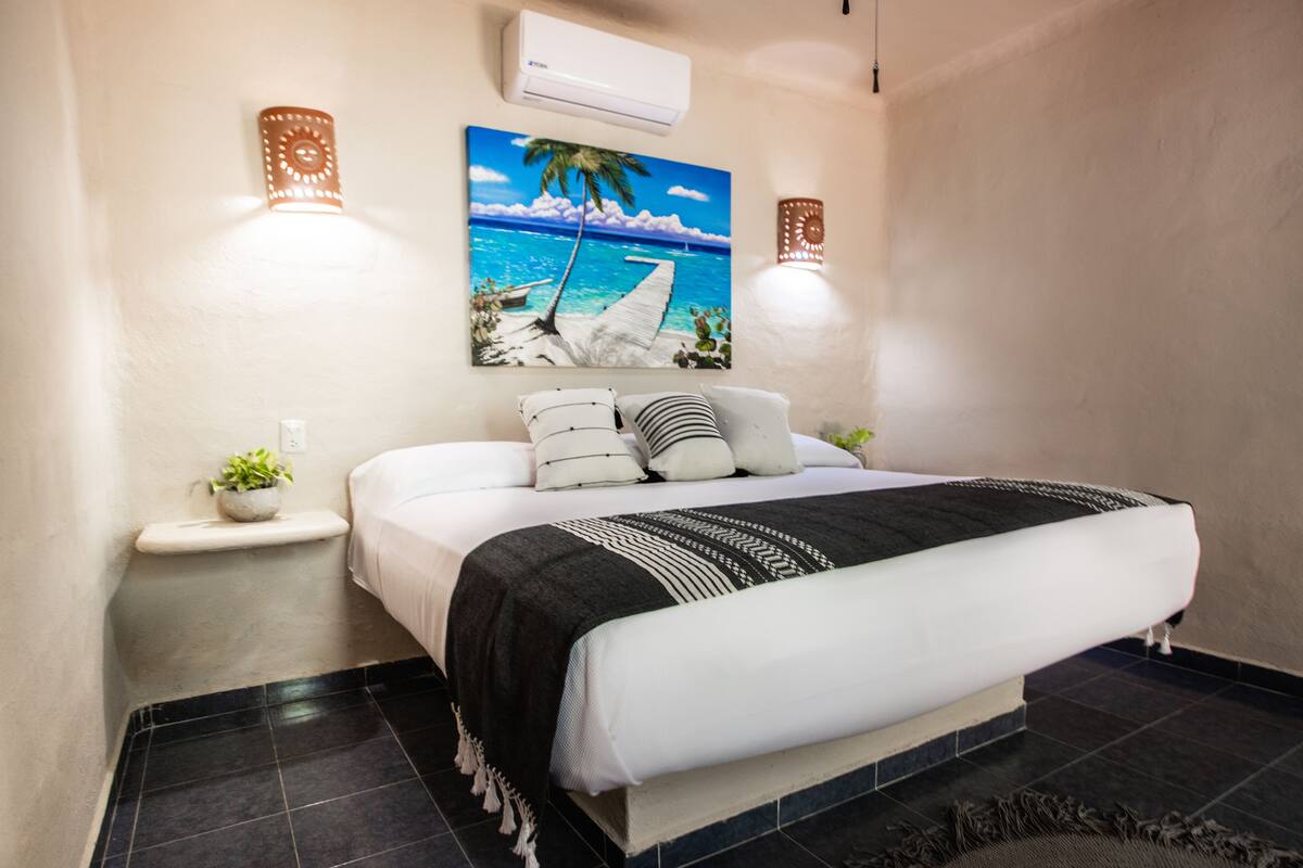 Een keurig opgemaakt slaapkamer met een bed, nachtkastjes, een wandairconditioner en een tropisch schilderij.