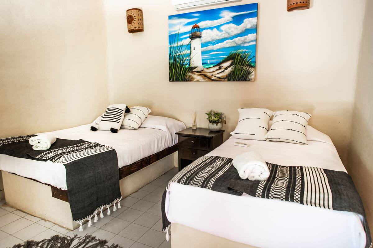 Een slaapkamer met twee bedden, elk voorzien van een bed met een decoratief kleed en kussens, een klein nachtkastje met een plant erop, en een schilderij van een vuurtoren aan de muur.