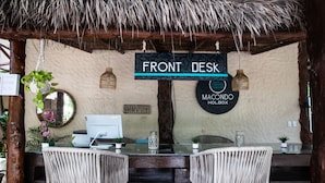 Un comptoir d’accueil avec une enseigne indiquant « RÉCEPTION » et « MACCONDO HOLBOX ». On y trouve deux fauteuils en osier et une table en verre, placés devant un ordinateur et un miroir.
