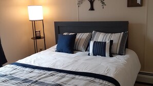 Una cama bien hecha con una colcha a rayas y varios cojines, una lámpara de noche y detalles decorativos en la pared, incluyendo un árbol y espejos enmarcados.