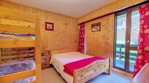 A wooden room with bunk beds, a single bed with a red blanket, a nightstand, and a window with curtains.