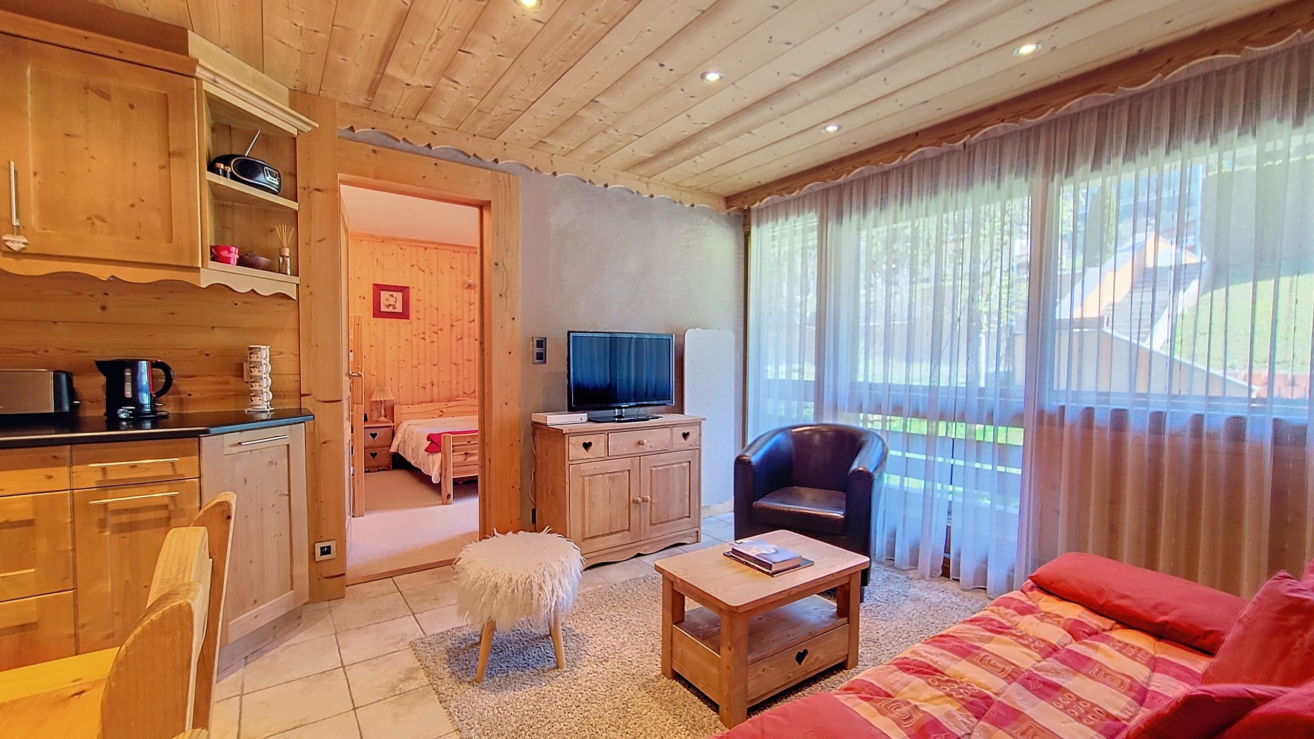 Una sala de estar acogedora con televisión, un sofá, una mesa de centro y una silla. Cuenta con una zona de cocina con armarios y un hervidor.