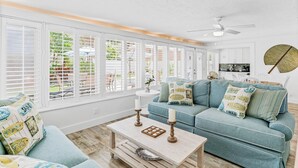 A spacious living room with a blue sofa, white walls, and large windows with blinds.