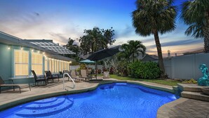 A swimming pool with lounge chairs and umbrellas, surrounded by palm trees and a building with string lights.