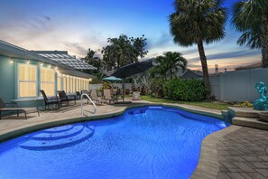 A swimming pool with lounge chairs and umbrellas, surrounded by palm trees and a building with string lights.