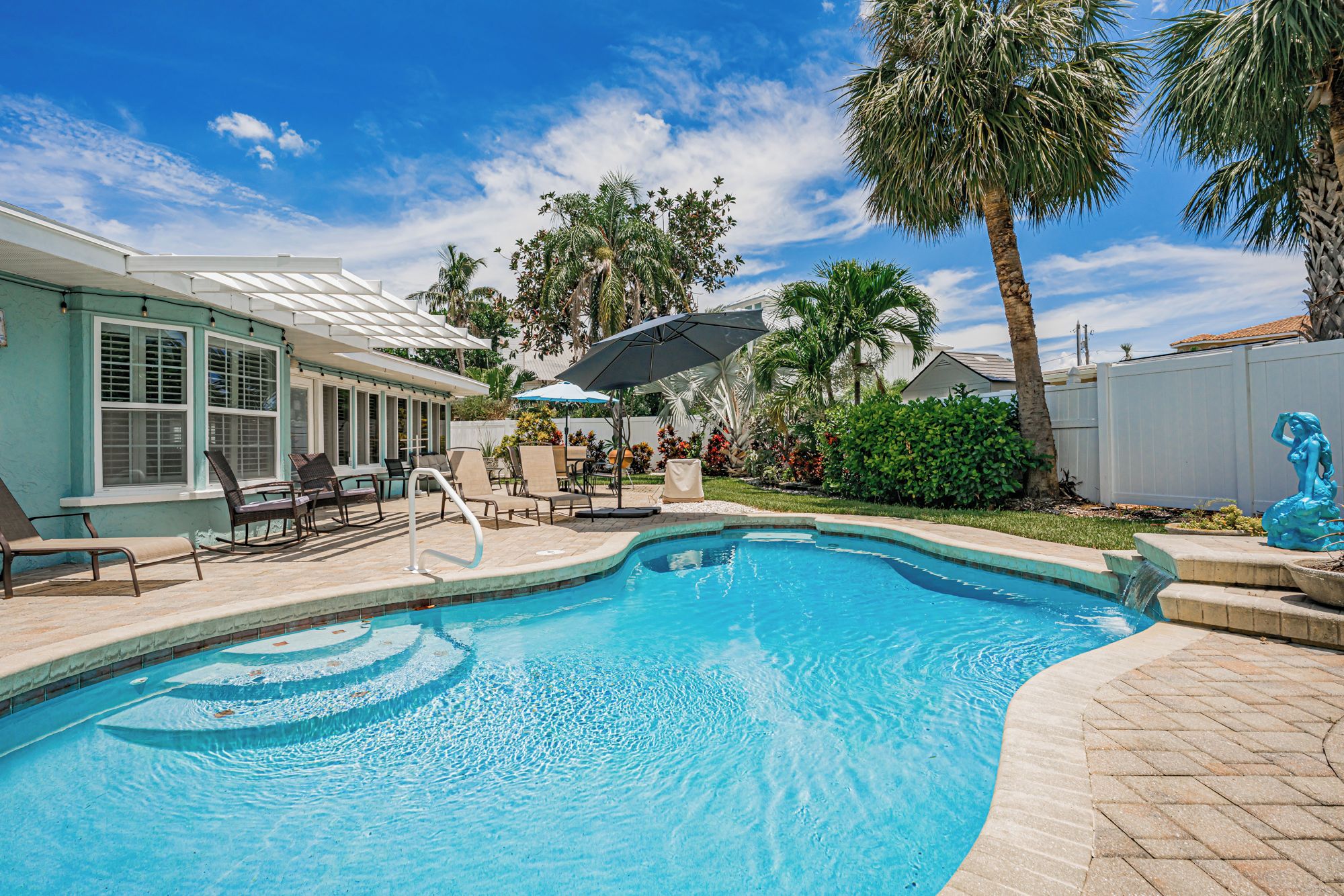 A swimming pool with lounge chairs and umbrellas, surrounded by palm trees and a house with a covered patio.