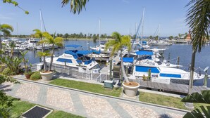 A marina with boats docked, a paved walkway, and palm trees.