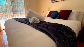 A neatly made bed with a white duvet, a dark blanket, and a rolled white towel. There are red and blue pillows on the bed.