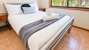 A neatly made bed with a white comforter, a grey blanket, and a rolled white towel. There is a wooden nightstand with a drawer, a window with a view of trees, and a metal bed frame.