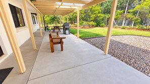 A covered patio area with a concrete floor, wooden benches, a grill, and a landscaped garden with a fence.