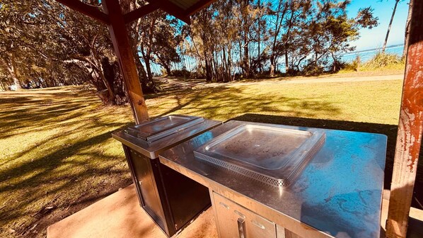 An outdoor kitchen with a sink and a grill, surrounded by trees and a clear sky.