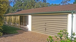 A single-story building with a garage door, a covered patio area, and a gravel driveway. - Home Away in Callala Bay (Callala Bay)