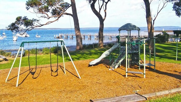 A playground with swings and a slide, located near a body of water with boats and a pier in the background. - Home Away in Callala Bay (Callala Bay)
