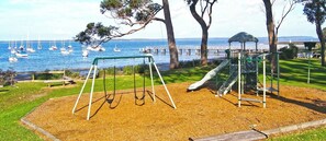 A playground with swings and a slide, located near a body of water with boats and a pier in the background. - Home Away in Callala Bay (Callala Bay)