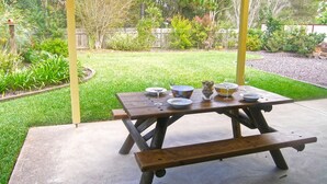 A wooden picnic table with benches, set on a concrete patio with a grassy yard and a wooden fence in the background. - Home Away in Callala Bay (Callala Bay)