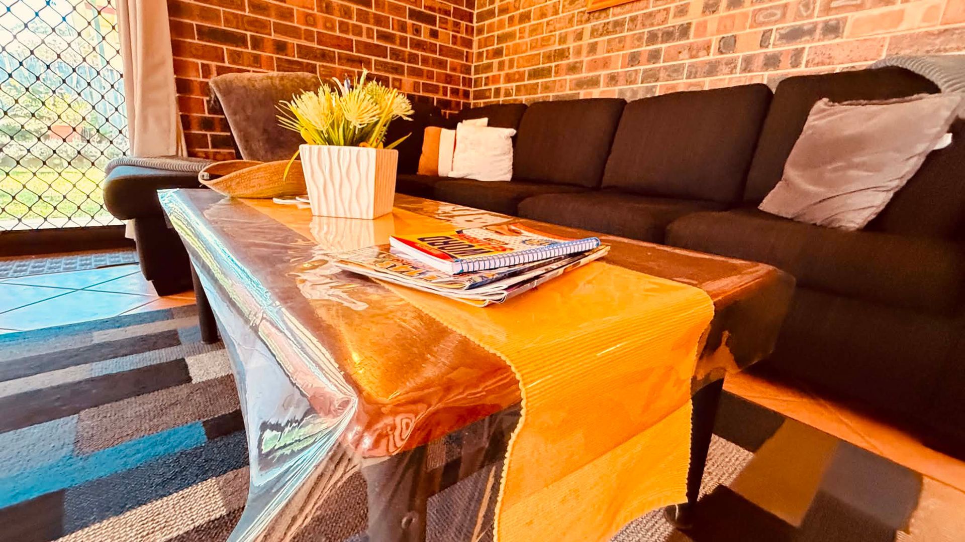 A living room with a sofa, a coffee table with magazines, a potted plant, and a brick wall.