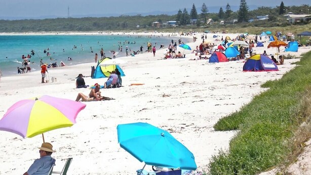Ein Strand mit Zelten und Sonnenschirmen, Menschen, die sich sonnen und schwimmen, und freier Blick auf das Meer.